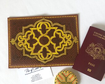 May include: A brown leather-bound journal with a gold geometric design. The journal is decorated with small gold beads. The journal is next to a passport and a small round decorative object.