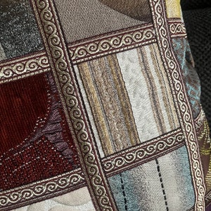 May include: A decorative cushion with a patchwork design featuring various shades of brown, beige, red, and blue. The cushion has a gold trim with a repeating scroll pattern.