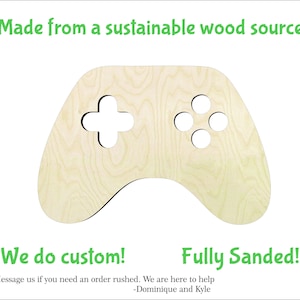 May include: A wooden controller-shaped cutout with a light wood grain finish. The controller features a directional pad and four circular buttons. Text at the top reads "Made from a sustainable wood source." Additional text includes "We do custom!" and "Fully Sanded!"