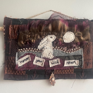 May include: Textile wall hanging with a white rabbit, a stitched moon, and the words "Honor your Wild Heart." The artwork uses a patchwork design in brown, burgundy, and black. A copper leaf charm hangs below.