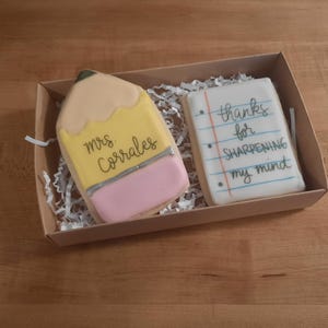 May include: Two decorated sugar cookies in a brown box with shredded paper. One cookie is shaped like a yellow pencil with a pink eraser and says "mrs. Corrales". The other cookie is shaped like a sheet of paper with lines and says "thanks for sharpening my mind".