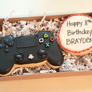 May include: A brown box filled with white shredded paper contains two decorated sugar cookies. One cookie is shaped like a black PlayStation controller with green, red, and white buttons. The other cookie is round and white with red icing around the edge and the text "Happy 8th Birthday BRAYDEN!" in black icing.