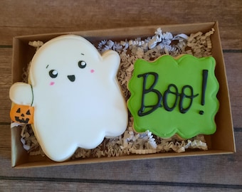 May include: A set of two Halloween-themed sugar cookies. One cookie is shaped like a ghost with a friendly face and a small pumpkin in its hand. The other cookie is green and has the word "Boo!" written in black icing.