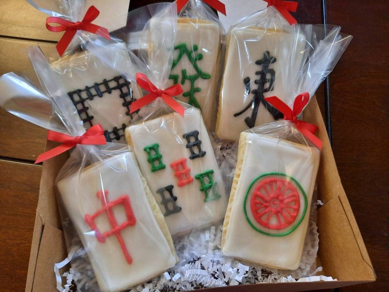 Puede incluir: Una caja de galletas decoradas, cada una envuelta individualmente y atada con una cinta roja. Las galletas presentan varios dise&ntilde;os, incluyendo caracteres chinos y patrones geom&eacute;tricos, en glaseado rojo, verde y negro.