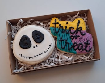 May include: Two decorated sugar cookies in a brown box with shredded paper. One cookie is a white Jack Skellington face and the other is a multi-colored patchwork design with the words "trick or treat" in black icing.