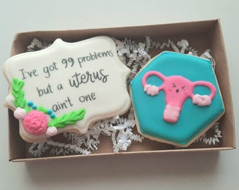 May include: Two decorated sugar cookies in a brown box with white shredded paper. One cookie is white with green and pink icing and the text "I've got 99 problems but a uterus ain't one". The other cookie is blue with a pink uterus with a cartoon face.