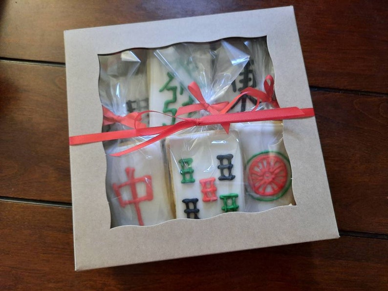 Puede incluir: Una caja de regalo con una ventana transparente que contiene varias galletas blancas decoradas con dise&ntilde;os coloridos. Las galletas est&aacute;n envueltas en celof&aacute;n transparente y atadas con una cinta roja. Los dise&ntilde;os incluyen caracteres chinos y una rueda roja.