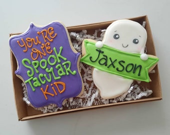 Puede incluir: Dos galletas de azúcar decoradas en una caja marrón. Una galleta es morada con letras verdes y naranjas que dicen "You're one Spooktacular Kid". La otra galleta es blanca con una pancarta verde que dice "Jaxson" y una cara de fantasma.