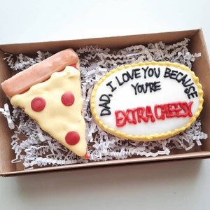 May include: A decorated cookie shaped like a pizza slice with red and white icing. Next to it is a round cookie with white icing and the text "Dad I love you because you're extra cheesy".
