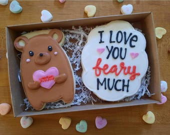 May include: A gift box with two decorated cookies. One cookie is brown and shaped like a bear holding a pink heart with the word "You" on it. The other cookie is white with the words "I love you" in black and "beary much" in red.
