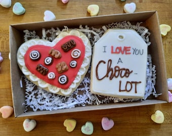 May include: A heart-shaped cookie decorated with red icing and chocolate candies, and a tag-shaped cookie with the text "I love you a Choco-Lot" in brown icing. Both cookies are in a brown box with white shredded paper.