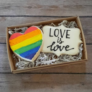 May include: A gift box filled with white paper shreds contains two decorated sugar cookies. One cookie is shaped like a heart with a rainbow design. The other cookie is a rectangular shape with the words "LOVE is love" written in black icing.