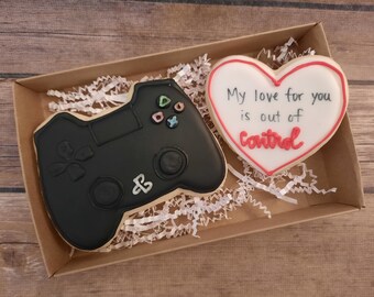 May include: A brown cardboard box with white tissue paper inside. The box contains two decorated cookies. One cookie is shaped like a video game controller and is black. The other cookie is shaped like a heart and is white with red lettering that says "My love for you is out of control."