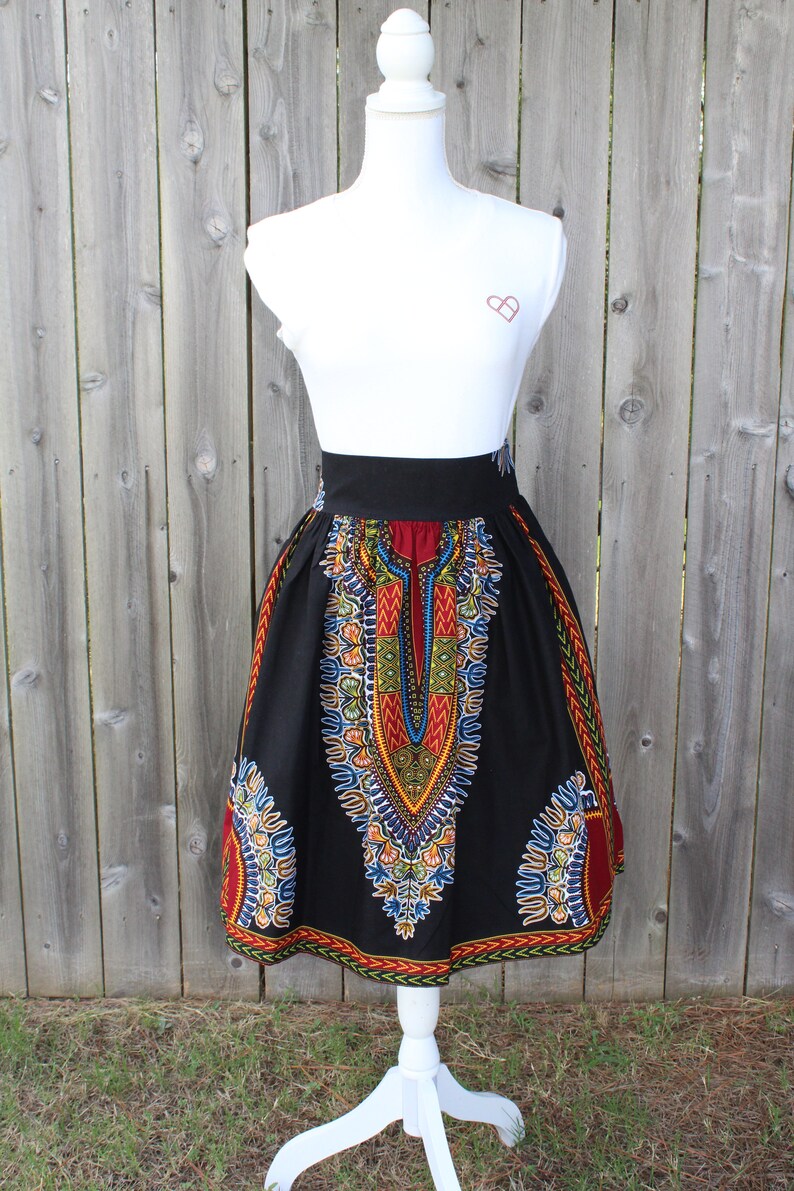 May include: A black skirt with a vibrant, colorful African print. The skirt features a wide black waistband and a flared silhouette. The print includes red, yellow, blue, and green geometric patterns. The skirt is displayed on a white mannequin.