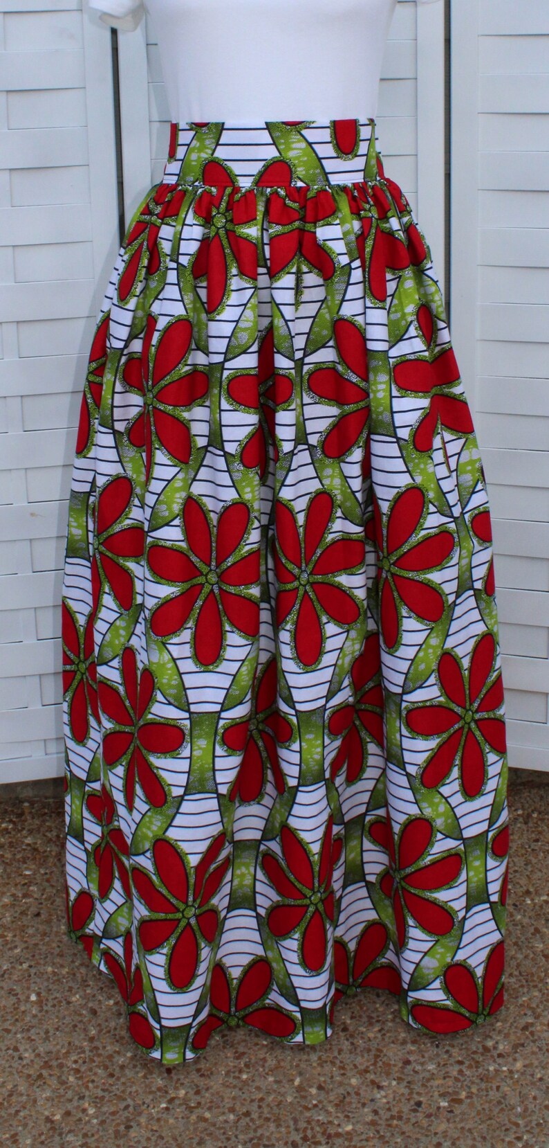 Peut inclure: Longue jupe pliss&eacute;e avec un motif floral vibrant. Le motif pr&eacute;sente de grandes fleurs rouges avec des feuilles vertes sur un fond blanc avec des rayures noires. La jupe est en tissu l&eacute;ger.
