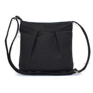 May include: A black crossbody bag with a long, adjustable strap. The bag is made of a solid, dark fabric with a simple, pleated design on the front. The strap is attached with silver-colored metal rings.