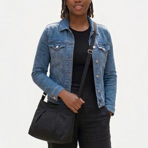 May include: A black crossbody bag with a pleated front, worn over the shoulder. The bag has a long, adjustable strap. The person is wearing a denim jacket and black pants.
