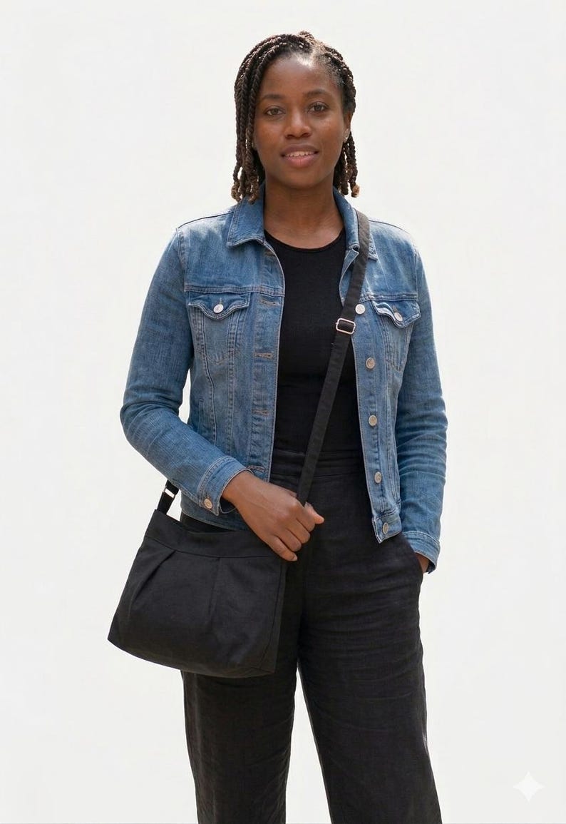 May include: A black crossbody bag with a pleated front, worn over the shoulder. The bag has a long, adjustable strap. The person is wearing a denim jacket and black pants.