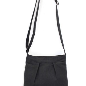 May include: A black crossbody bag with a long, adjustable strap. The bag features a rectangular shape with pleats at the bottom. The strap is attached to the bag with metal hardware. The bag is made of a solid black fabric.
