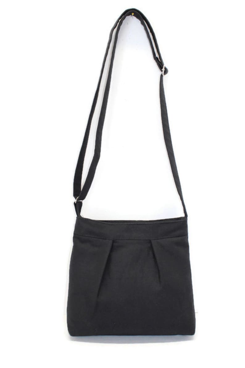 May include: A black crossbody bag with a long, adjustable strap. The bag features a rectangular shape with pleats at the bottom. The strap is attached to the bag with metal hardware. The bag is made of a solid black fabric.