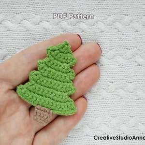 Op de afbeelding: Een groene gehaakte kerstboomversiering met een bruine gehaakte stam. De versiering wordt in een hand gehouden tegen een witte gebreide achtergrond. De tekst "PDF Pattern" is zichtbaar bovenaan de afbeelding.