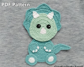 May include: A crocheted green and blue dinosaur with white accents. The dinosaur is sitting on a gray fabric background. The text "PDF Pattern" is visible at the top of the image.