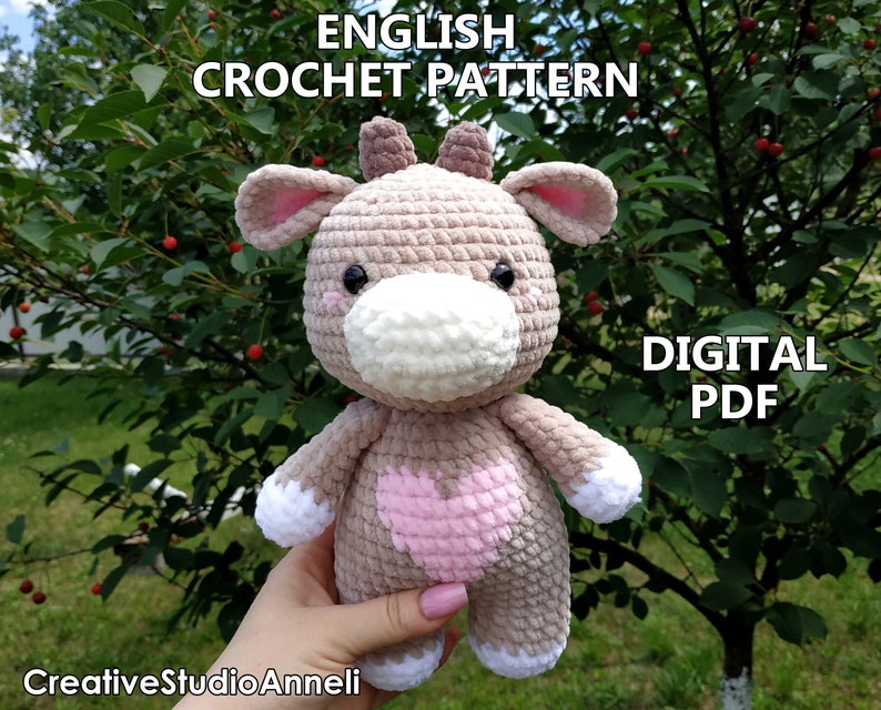 May include: Crochet pattern for a plush brown and pink cow toy with a white face. The pattern is available as a digital PDF download. The text "ENGLISH CROCHET PATTERN" and "DIGITAL PDF" is visible in the image.