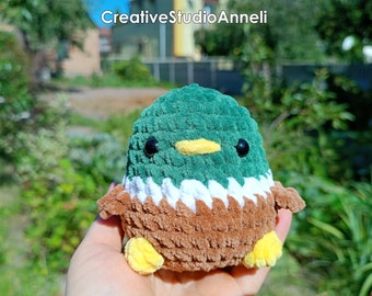 Puede incluir: Un pequeño juguete de peluche de pato tejido a crochet con hilo verde, marrón, blanco y amarillo. El pato tiene ojos negros y un pico amarillo.