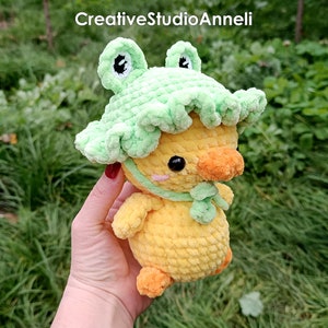 K&ouml;nnte beinhalten: Eine gelbe geh&auml;kelte Ente, die einen gr&uuml;nen geh&auml;kelten Froschhut tr&auml;gt. Die Ente hat einen kleinen orangefarbenen Schnabel und schwarze Augen. Der Froschhut hat gr&uuml;ne Augen und einen gr&uuml;nen K&ouml;rper.