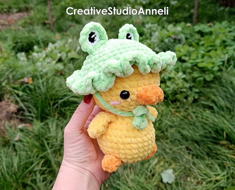 K&ouml;nnte beinhalten: Eine gelbe geh&auml;kelte Ente, die einen gr&uuml;nen geh&auml;kelten Froschhut tr&auml;gt. Die Ente hat einen kleinen orangefarbenen Schnabel und schwarze Augen. Der Froschhut hat gr&uuml;ne Augen und einen gr&uuml;nen K&ouml;rper.