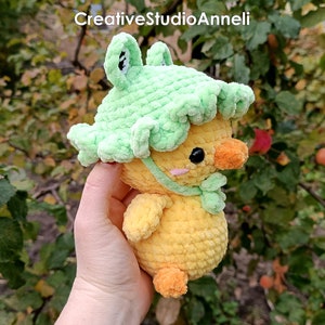 K&ouml;nnte beinhalten: Ein gelbes H&auml;kel-K&uuml;kenspielzeug, das einen gr&uuml;nen H&auml;kel-Froschhut tr&auml;gt. Das K&uuml;ken hat einen kleinen orangefarbenen Schnabel und schwarze Augen. Das Spielzeug wird in einer Hand vor einem gr&uuml;nen, belaubten Hintergrund gehalten.