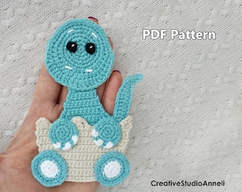 May include: Crochet pattern for a teal dinosaur sitting in a beige egg. The pattern is available as a PDF download. The text "PDF Pattern" is visible in the image.