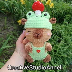 May include: A handmade crochet llama doll wearing a light green frog hat and overalls with a red heart. A red mushroom sits atop the hat. The doll has closed eyes and pink cheeks. The text "CreativeStudioAnneli" is at the bottom.