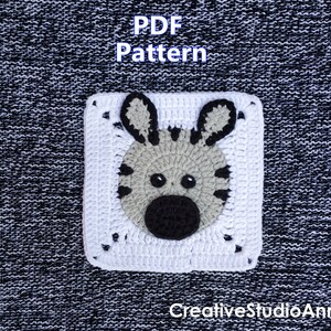Pode incluir: Um quadrado branco de crochê com um rosto de zebra com detalhes em preto e cinza. O texto "PDF Pattern" está no topo e "CreativeStudioAnneli" na parte inferior. O fundo é um tecido texturizado em preto e branco.