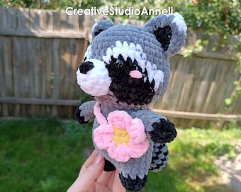 Puede incluir: Un juguete de peluche de mapache tejido a crochet con una flor rosa en el pecho. El juguete es gris, negro y blanco con ojos negros y una nariz rosa.