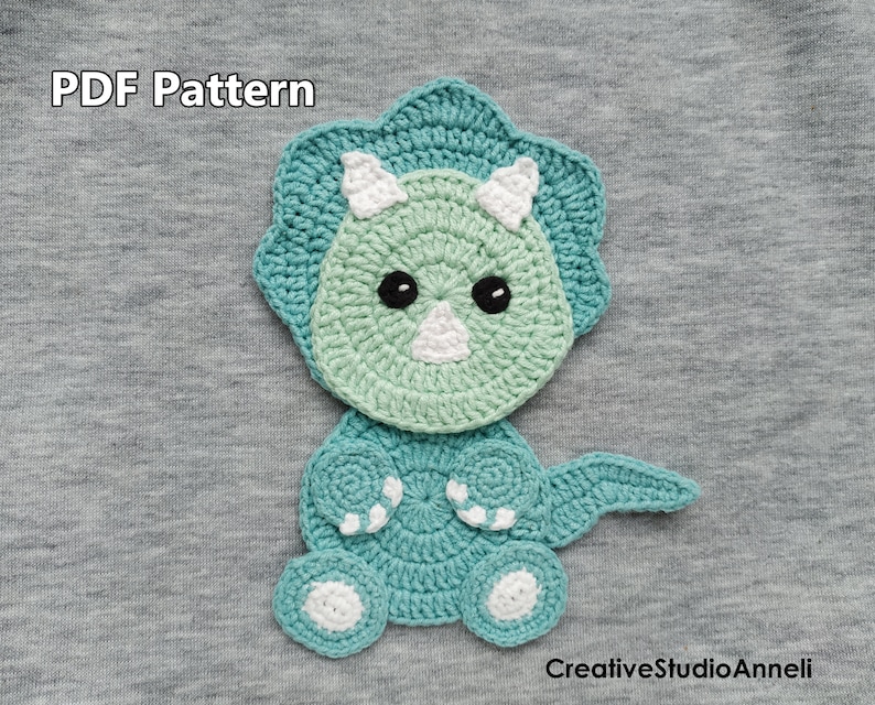 May include: A crocheted green and blue dinosaur with white accents. The dinosaur is sitting on a grey fabric background. The text "PDF Pattern" is visible at the top of the image. The text "CreativeStudioAnnelli" is visible at the bottom of the image.