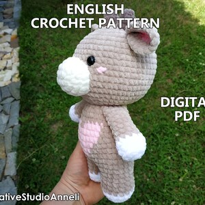 May include: Crochet pattern for a plush toy in the shape of a brown bear with a pink heart on its chest. The pattern is available as a digital PDF download. The text "ENGLISH CROCHET PATTERN" and "DIGITAL PDF" is visible in the image.