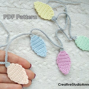 Puede incluir: Una cadena de bombillas de luz de ganchillo en colores pastel, incluyendo rosa, azul, amarillo y melocotón. La cadena está hecha con hilo gris y se muestra sobre una superficie de punto blanco. El texto "PDF Pattern" es visible en la imagen.