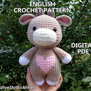 May include: Crochet pattern for a plush grey and pink cow with a white nose and heart on its chest. The pattern is available as a digital PDF download. The text "ENGLISH CROCHET PATTERN" and "DIGITAL PDF" is visible in the image.