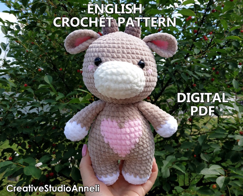 May include: Crochet pattern for a plush grey and pink cow with a white nose and heart on its chest. The pattern is available as a digital PDF download. The text "ENGLISH CROCHET PATTERN" and "DIGITAL PDF" is visible in the image.