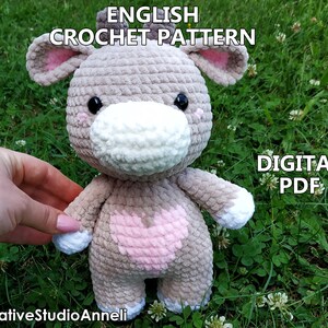 May include: Crochet pattern for a plush toy cow in beige, pink, and white. The toy has a heart on its chest and a white nose. The pattern is available as a digital PDF download. The text "ENGLISH CROCHET PATTERN" and "DIGITAL PDF" is visible in the image.