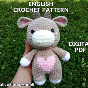 May include: Crochet pattern for a plush toy cow with a pink heart on its chest. The cow is gray with white accents and black eyes. The pattern is available as a digital PDF download. The text "ENGLISH CROCHET PATTERN" and "DIGITAL PDF" is visible in the image.