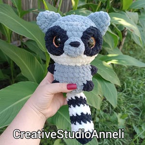 May include: A handmade crochet raccoon plush toy in shades of gray, black, and white. The raccoon has large, expressive eyes and a striped tail. The text "CreativeStudioAnneli" is visible at the bottom of the image.