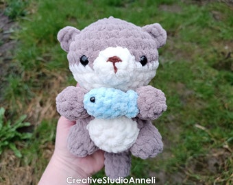Puede incluir: Una nutria de crochet gris y blanco sosteniendo un pez de crochet azul claro. La nutria tiene ojos negros y una nariz marrón.