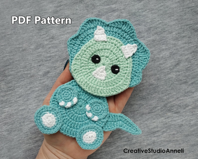 May include: Crocheted amigurumi triceratops pattern, PDF pattern, green and blue, with white accents, "PDF Pattern" text.