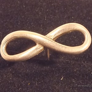 Puede incluir: Un pin de plata con forma de símbolo de infinito. El pin es un diseño simple con un acabado liso.