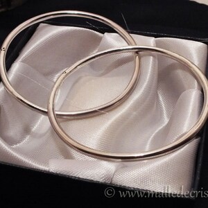 May include: Two silver bangle bracelets, one slightly larger than the other, are displayed on a white satin background inside a black box. The box has a label on the bottom that reads "www.malledecristal.fr".