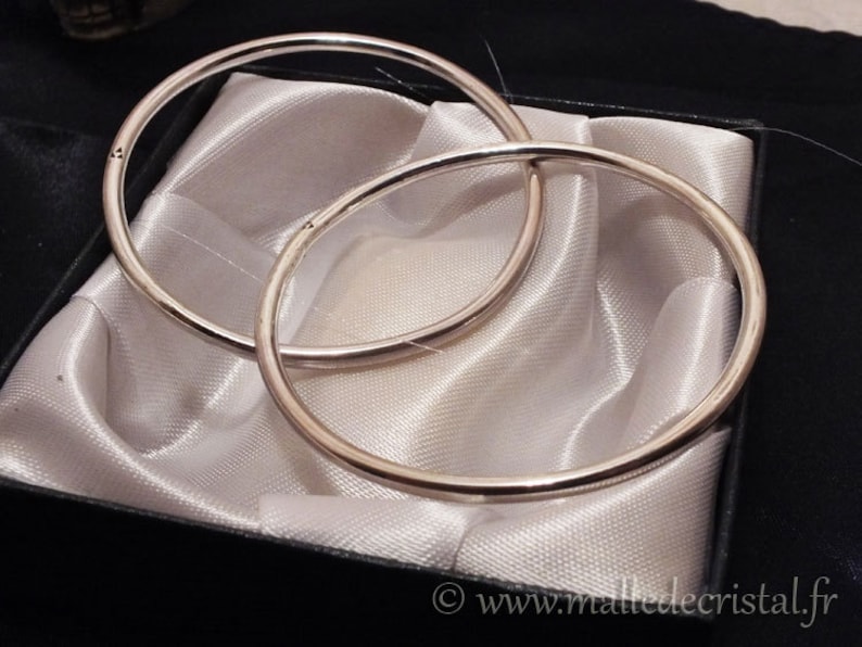 May include: Two silver bangle bracelets, one slightly larger than the other, are displayed on a white satin background inside a black box. The box has a label on the bottom that reads "www.malledecristal.fr".