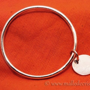 May include: A silver bangle bracelet with a round silver charm hanging from it. The bracelet is on a red fabric background.