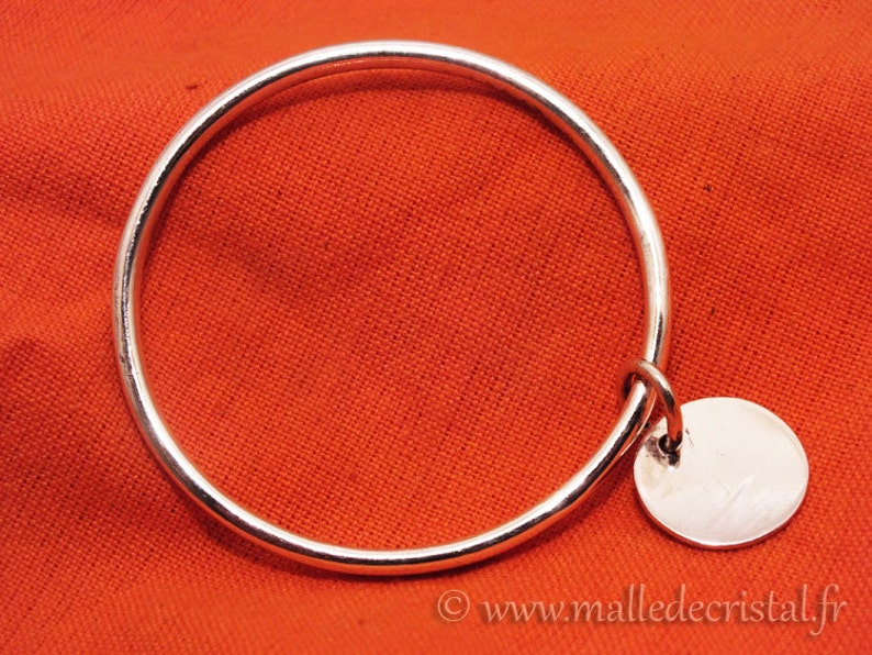 May include: A silver bangle bracelet with a round silver charm hanging from it. The bracelet is on a red fabric background.